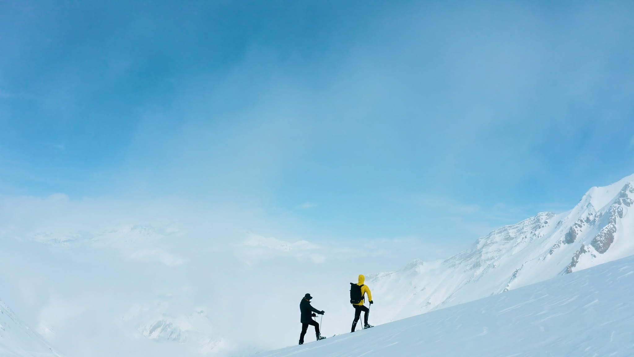 Two small hikers stand on a snowy mountain slope under a bright blue sky, with snow-covered peaks in the background.