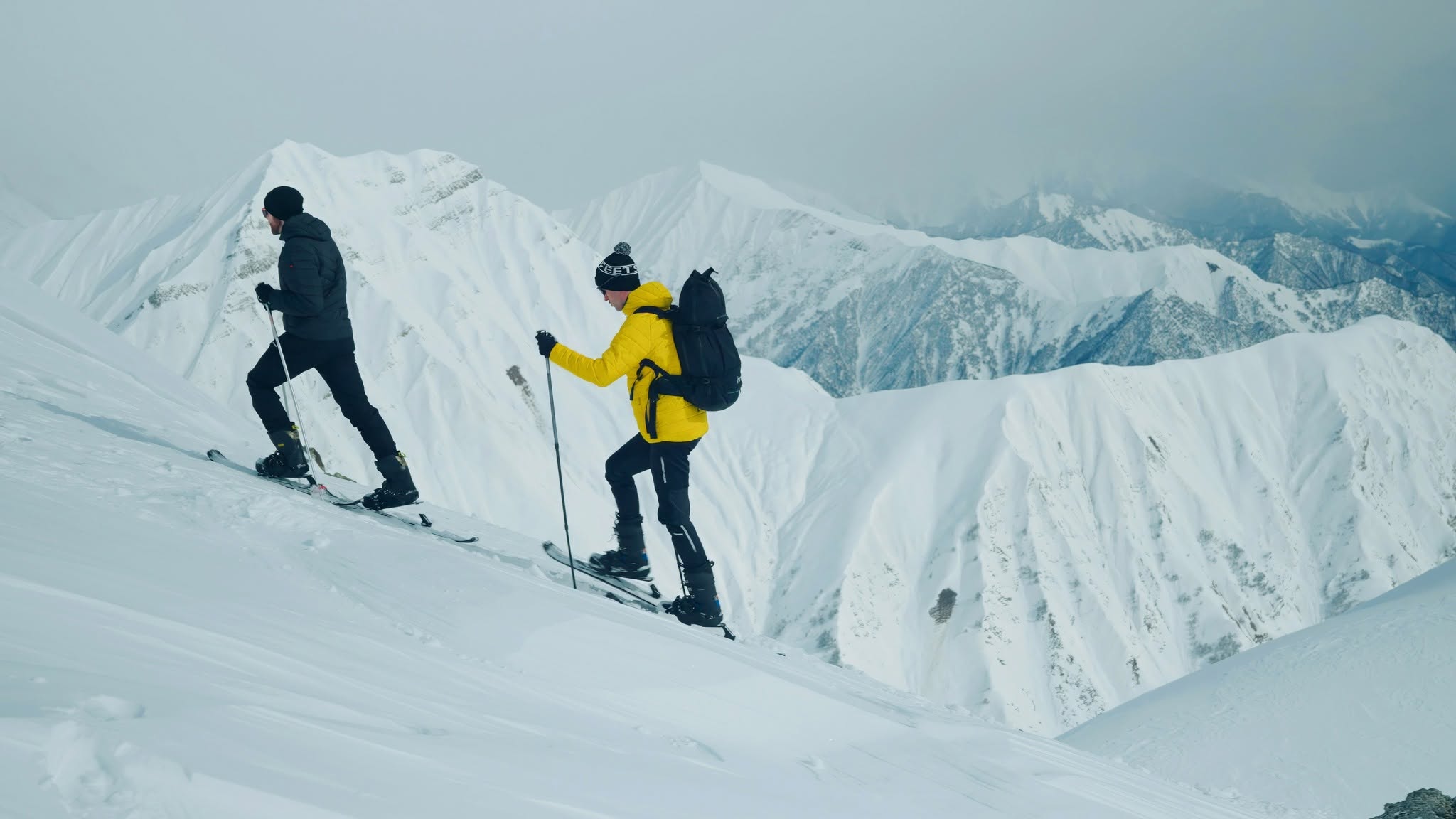 Two skiers traverse a snowy mountain slope with snow-covered peaks in the background under an overcast sky.