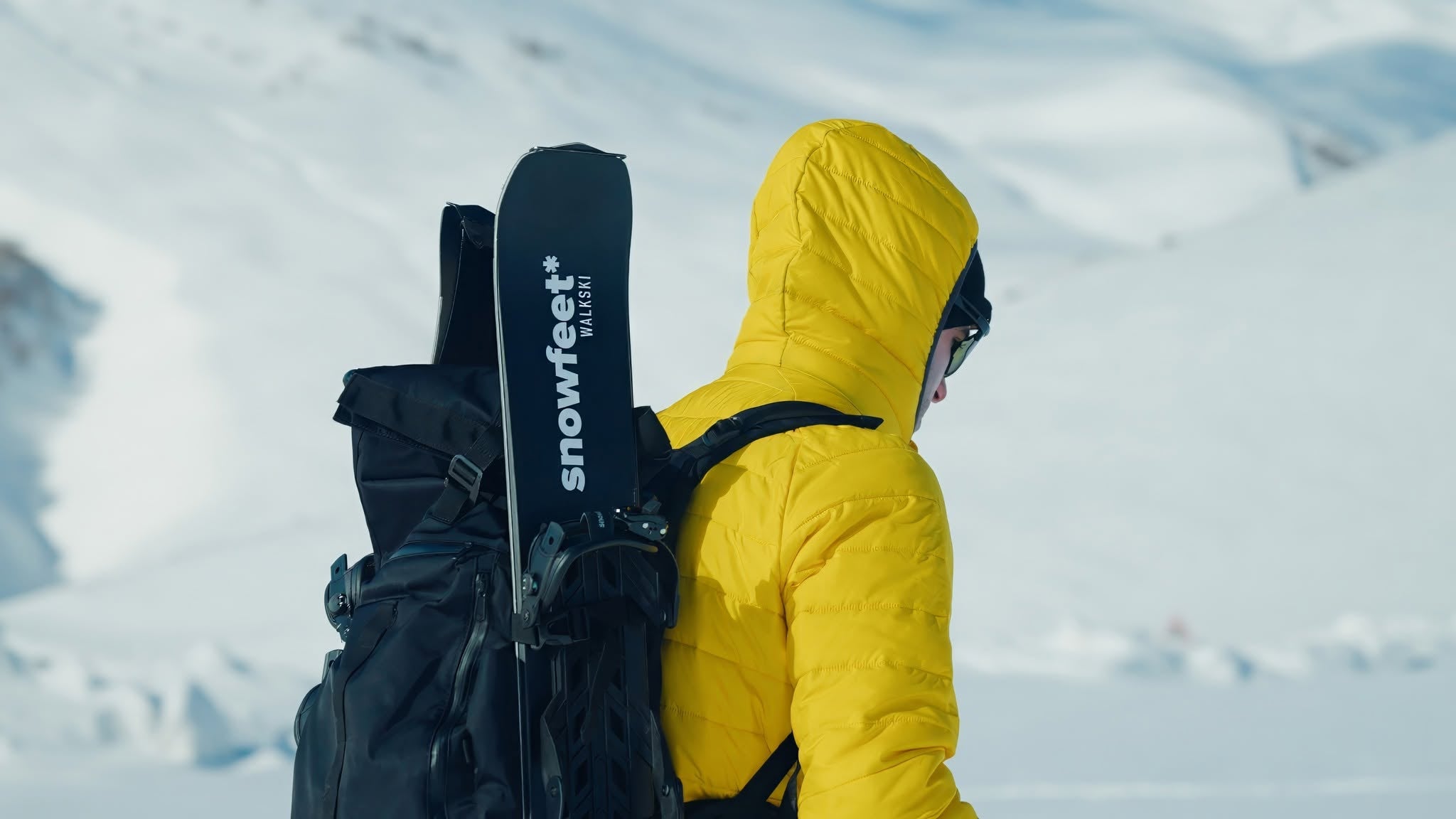 A person in a bright yellow hooded jacket stands in a snowy outdoor setting, carrying a black backpack with a black Snowfeet+ ski attached.